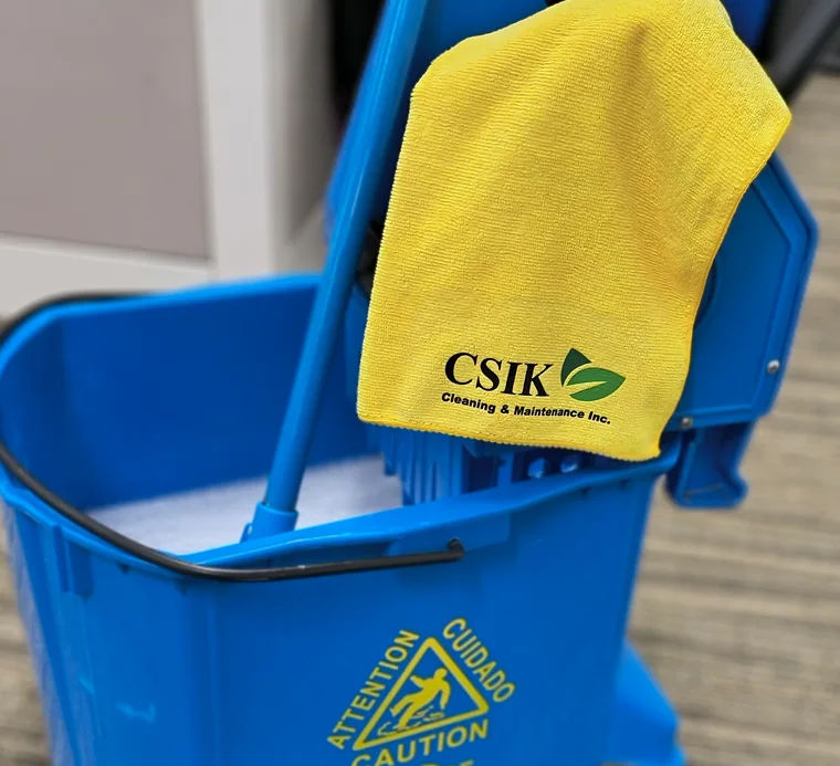 A close-up shot of a bright blue commercial mop bucket with a wringer attachment sitting on a patterned office carpet. A vibrant yellow microfiber towel branded with the C&K Cleaning & Maintenance Inc. logo hangs neatly over the side of the wringer mechanism. A yellow caution triangle sign reading "Attention / Cuidado / Caution" is visible on the front of the bucket, highlighting professional floor care equipment.