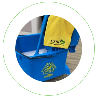 A close-up, circular-cropped shot of a bright blue commercial mop bucket with a wringer attachment sitting on a carpeted office floor. The bucket features a yellow "Caution / Cuidado / Attention" wet floor safety symbol on the front. A yellow microfiber cloth branded with the C&K Cleaning & Maintenance Inc. logo is draped neatly over the wringer handle, with a blue mop handle resting inside the bucket filled with soapy cleaning solution.