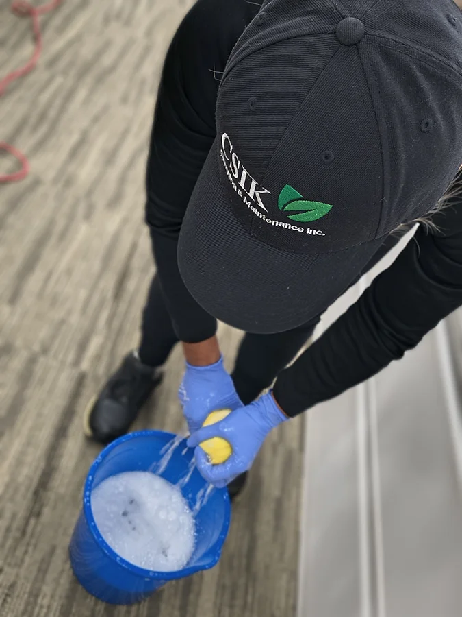 A top-down, bird's-eye view of a janitorial worker wearing a black baseball cap embroidered with the green and white CSIK Cleaning & Maintenance Inc. logo. The worker wears blue disposable nitrile gloves and wrings out a yellow cleaning sponge into a blue plastic bucket filled with foamy, sudsy water over patterned office carpet.