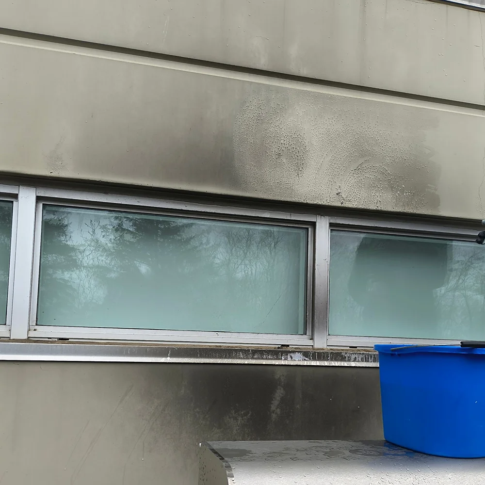 A close-up shot of a dirty commercial building exterior wall above windows showing heavy grime, dark soot buildup, and water marks prior to power washing, with a blue cleaning bucket positioned in the foreground.